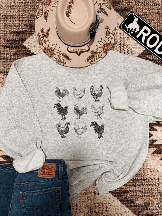 Flat lay of ash grey Chicken Crewneck featuring a hand drawn flock of roosters and hens, styled with denim jeans and a sunflower etched Hides & Kinds hat.