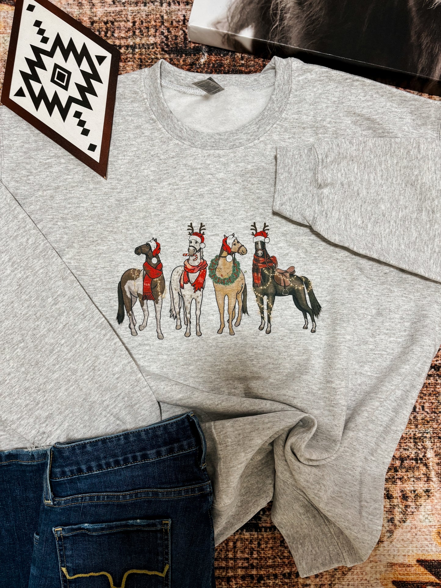 Christmas Horses crewneck sweatshirt in ash grey with five festive horses wearing western holiday gear, styled with jeans on a rustic background
