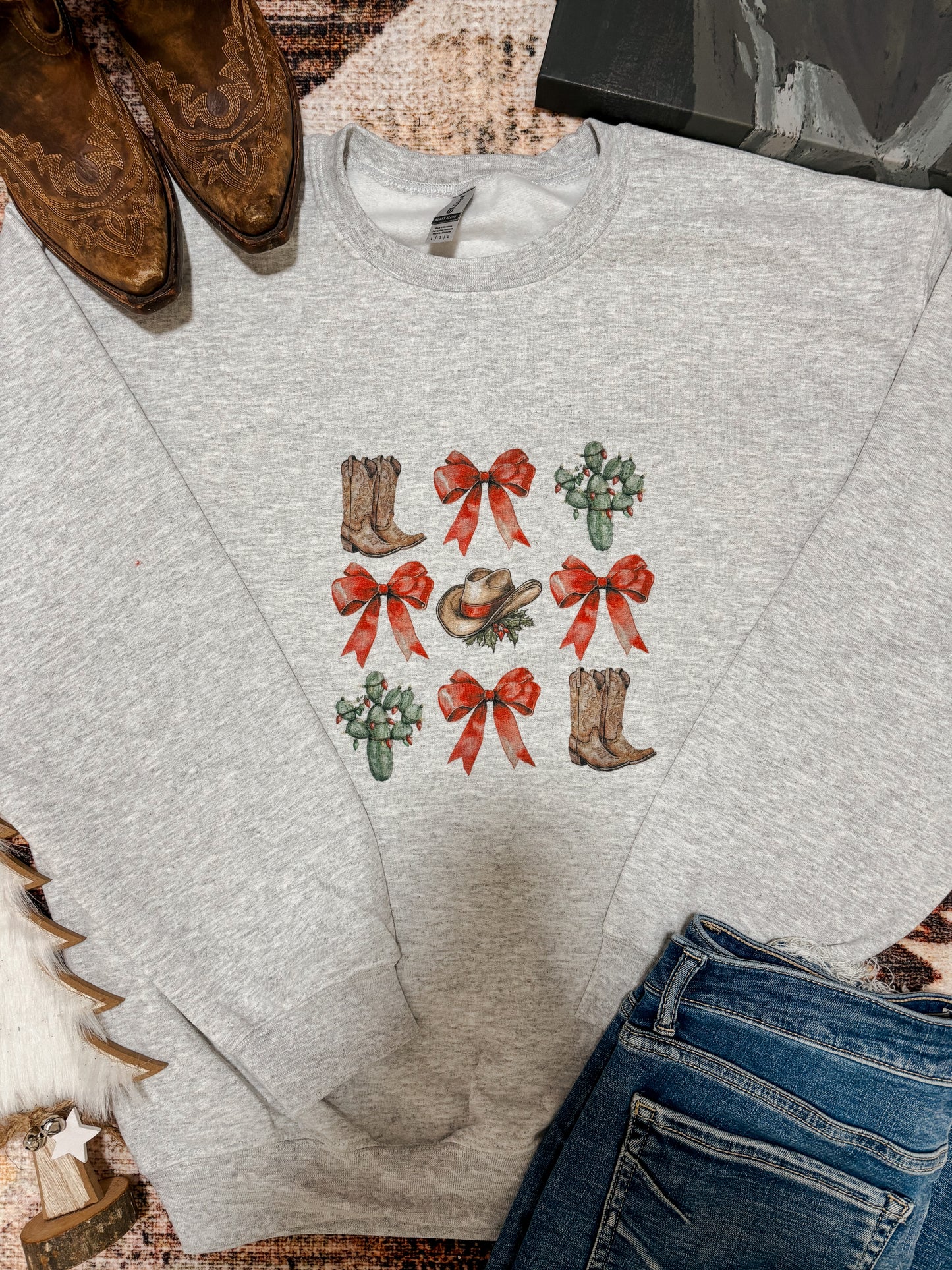 Grey crewneck with western Christmas graphics including red bows, cowboy boots, cactus, and cowboy hat, styled flat lay with jeans and boots.