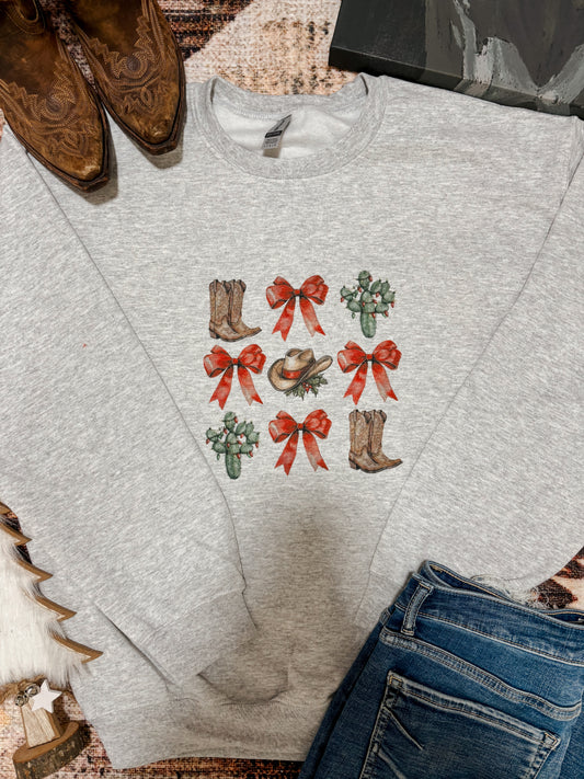 Grey crewneck with western Christmas graphics including red bows, cowboy boots, cactus, and cowboy hat, styled flat lay with jeans and boots.