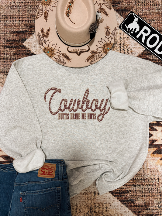 Grey crewneck with bold “Cowboy Butts Drive Me Nuts” rope text, styled with sunflower cowboy hat and Levi’s jeans on a western rug