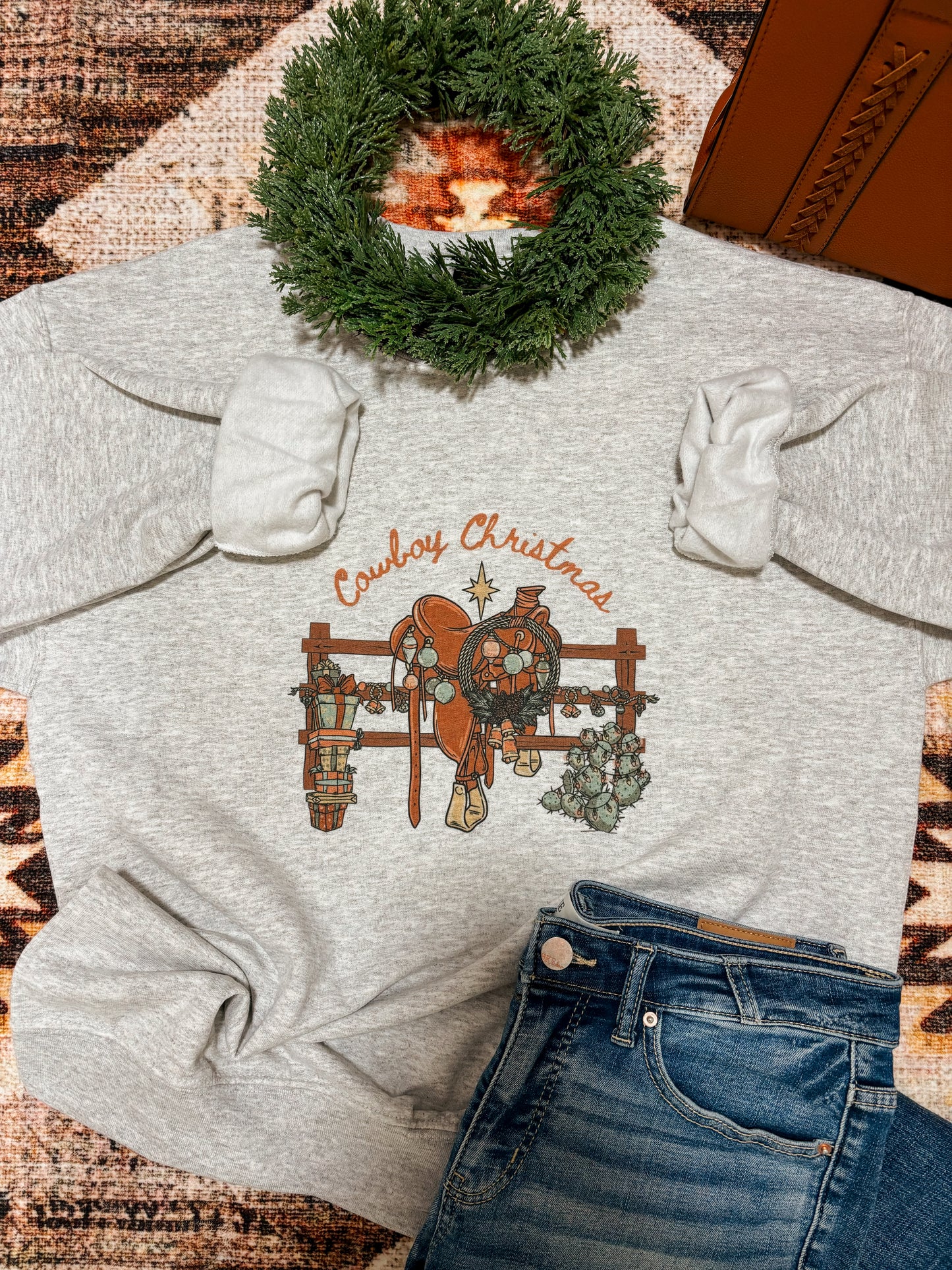 Ash grey Cowboy Christmas crewneck by Hides & Kinds styled with holiday wreath, jeans, and rustic western background