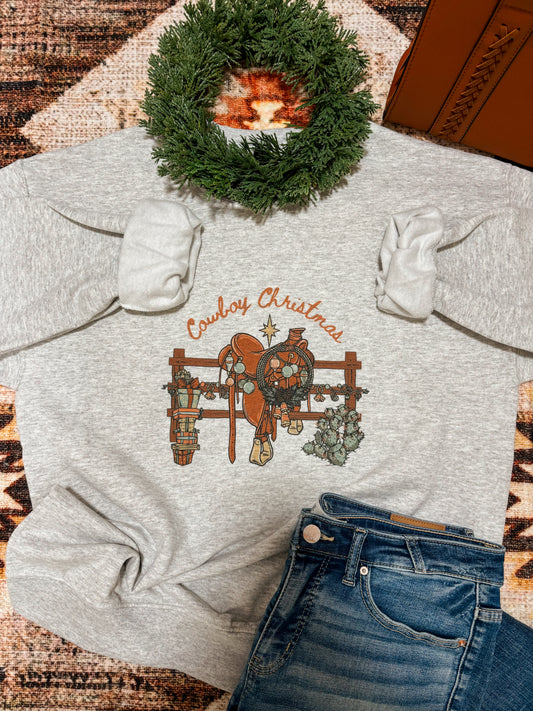 Ash grey Cowboy Christmas crewneck by Hides & Kinds styled with holiday wreath, jeans, and rustic western background