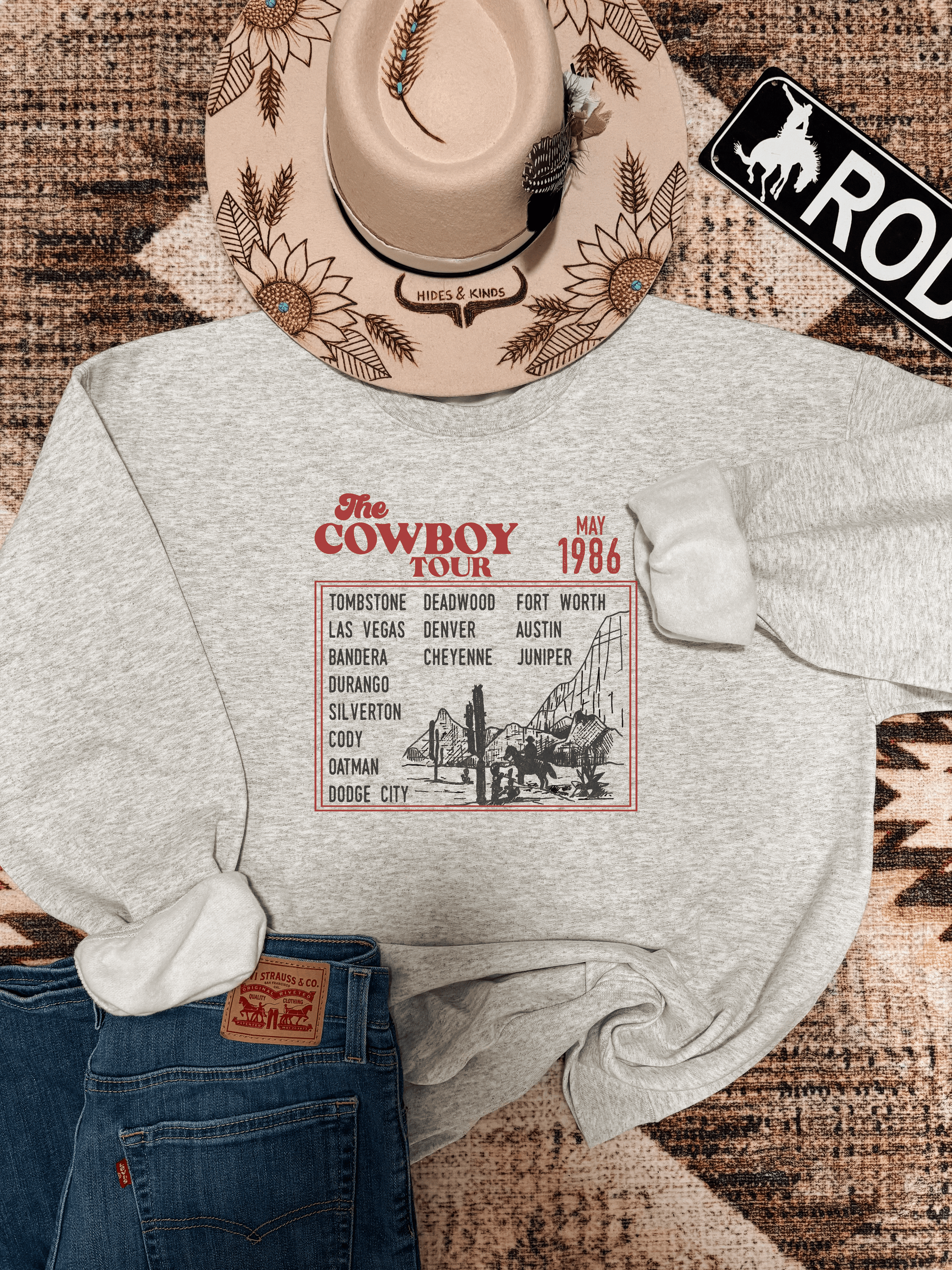 Ash grey Cowboy Tour crewneck from Hides & Kinds featuring vintage western cities graphic and rustic flat lay styling with hat and jeans