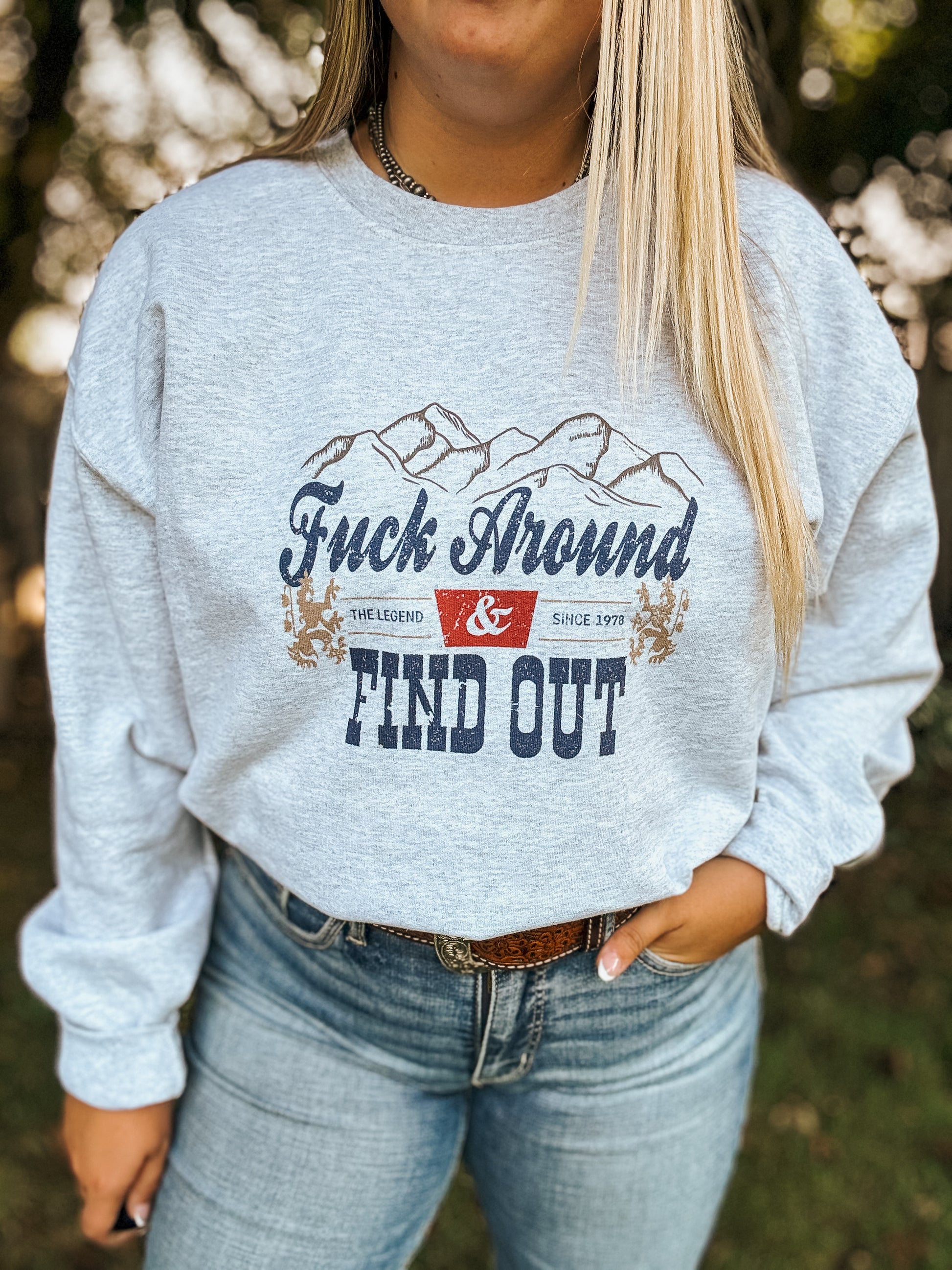 Woman wearing grey crewneck that says "Fuck Around & Find Out" with western mountain design