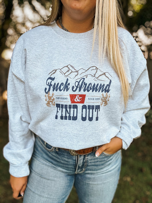 Woman wearing grey crewneck that says "Fuck Around & Find Out" with western mountain design