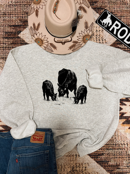 Flat lay of ash grey Herd It Crewneck featuring a black cattle herd graphic, styled with denim, sunflower Hides & Kinds hat, and western themed accents.