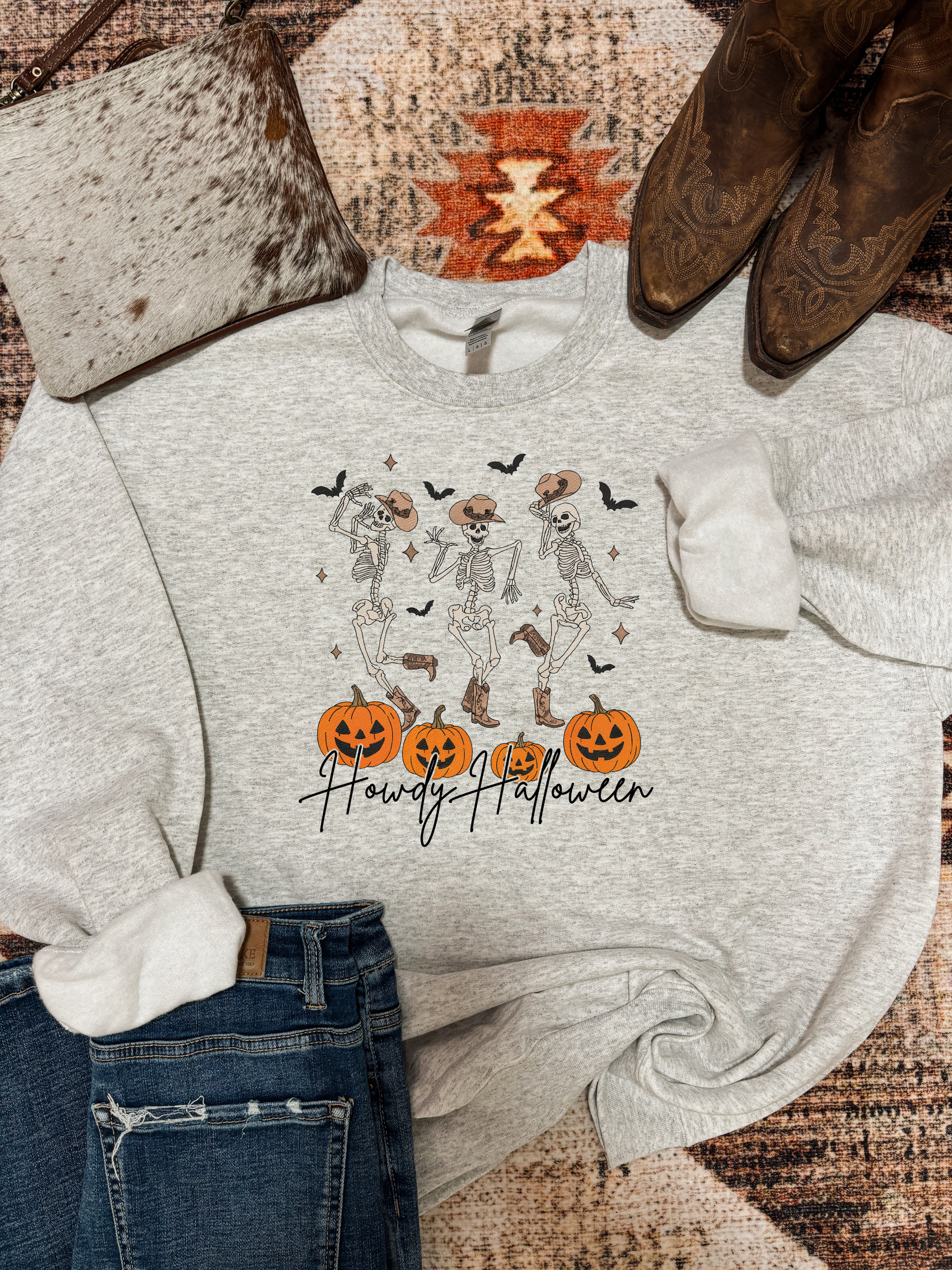 Light grey crewneck with three dancing skeletons in cowboy hats above carved pumpkins and “Howdy Halloween” text, styled with jeans, brown boots, and a cowhide purse