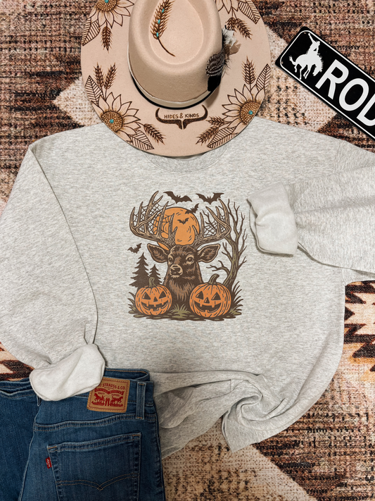 Ash grey crewneck featuring a buck with antlers wrapped in spiderwebs, flanked by jack-o’-lanterns and bats in a spooky fall forest scene, styled with jeans and a sunflower western hat