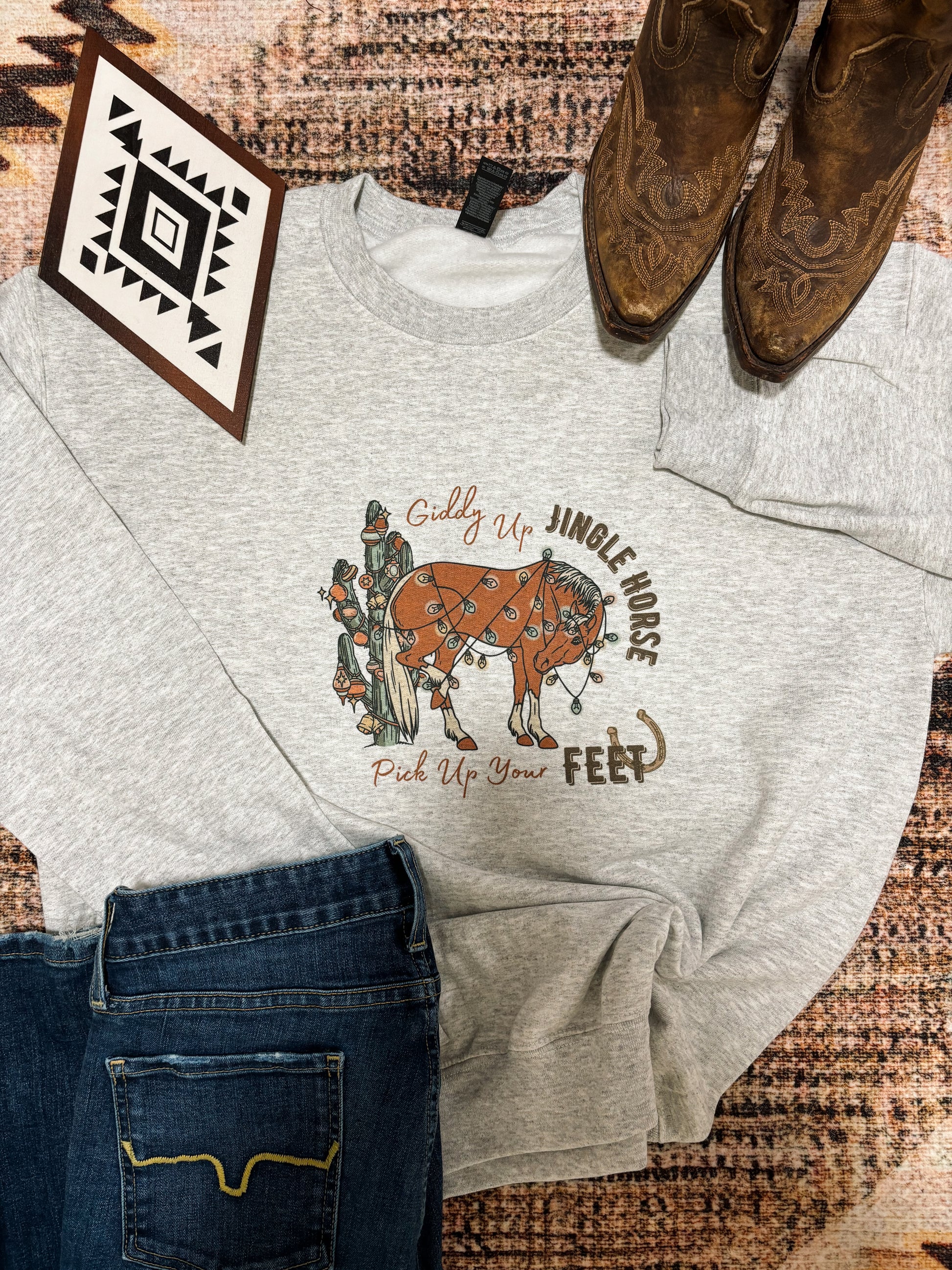 Hides and Kinds Jingle Horse crewneck sweatshirt laid out with cowboy boots, western decor, and denim jeans