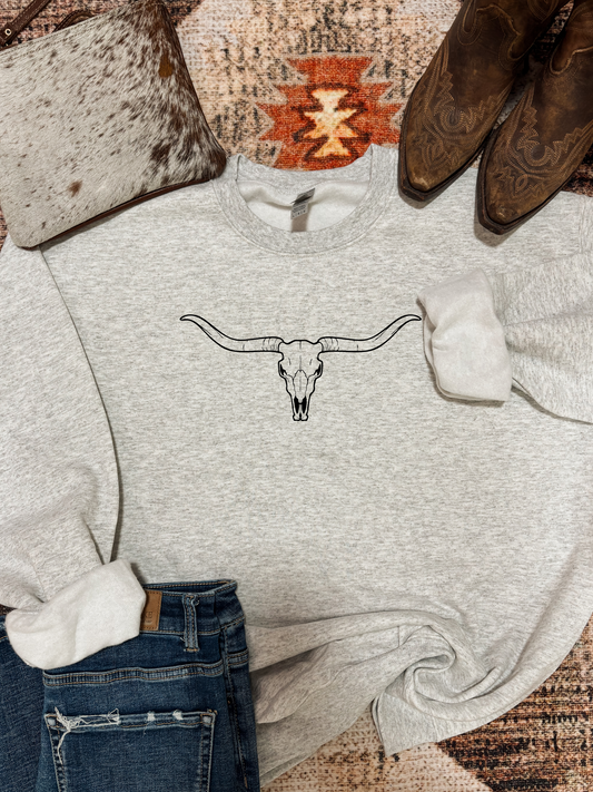 Ash grey crewneck with minimalist black longhorn skull graphic, styled flat lay with jeans, boots, and cowhide bag.