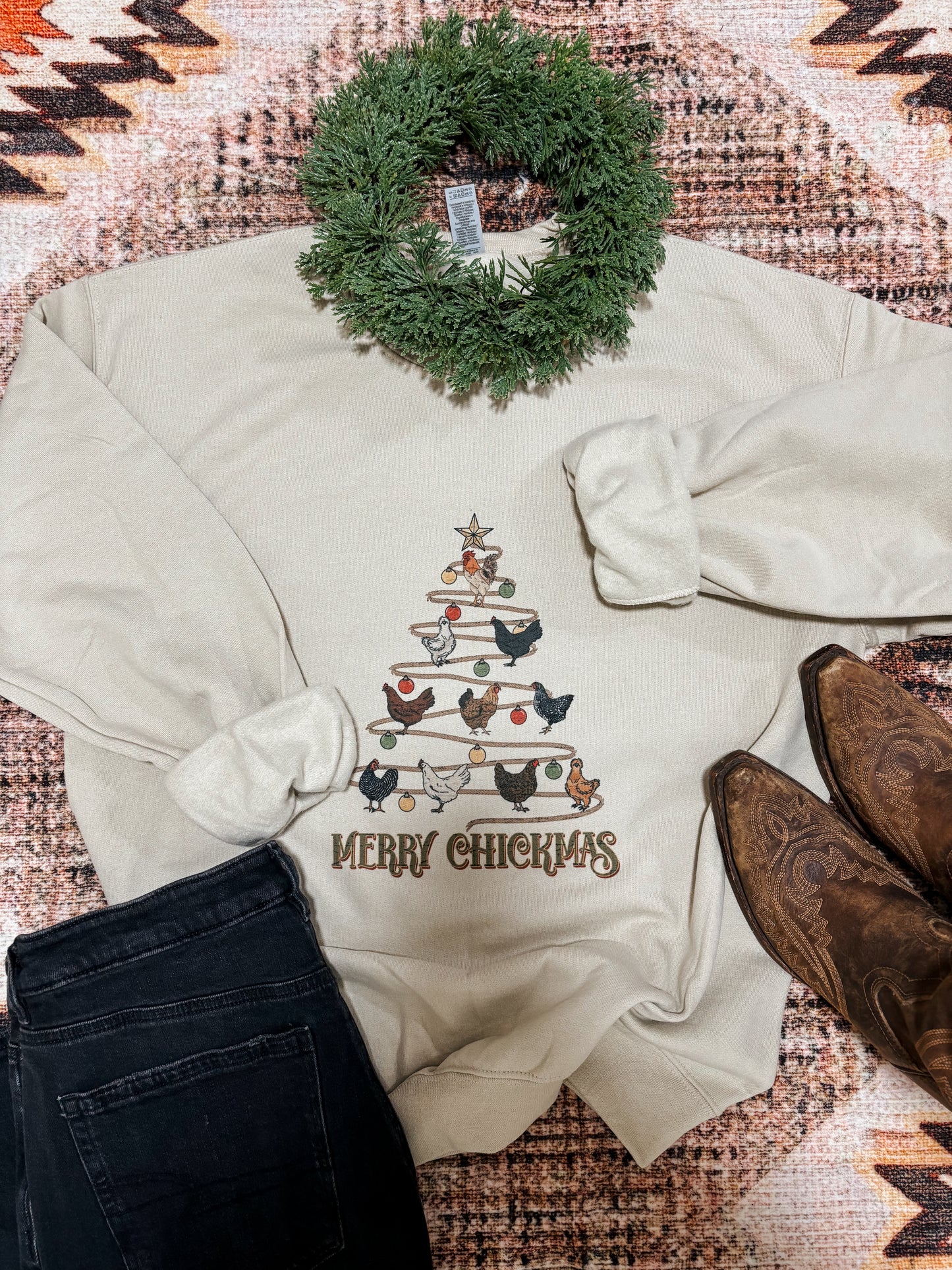 Flatlay of Merry Chickmas crewneck showing full chicken Christmas tree graphic with boots, garland, and denim styling
