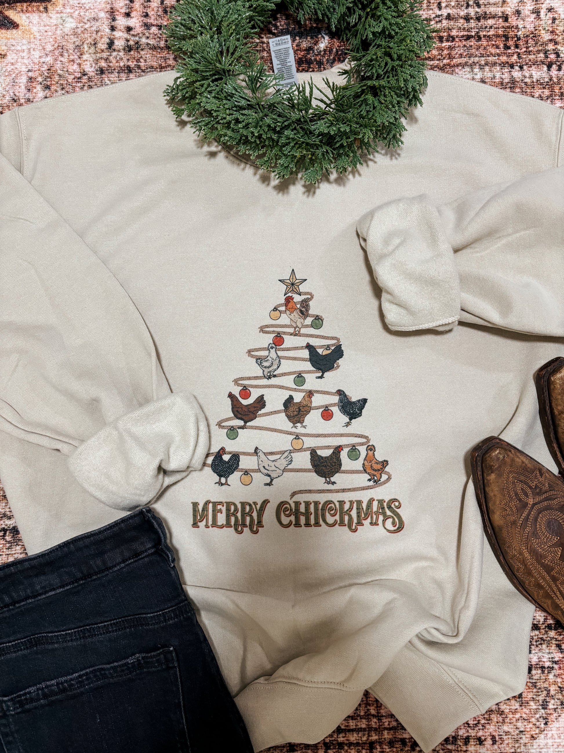 Cream crewneck with Christmas chicken tree design, styled with a green garland, black jeans, and cowboy boots on a western rug