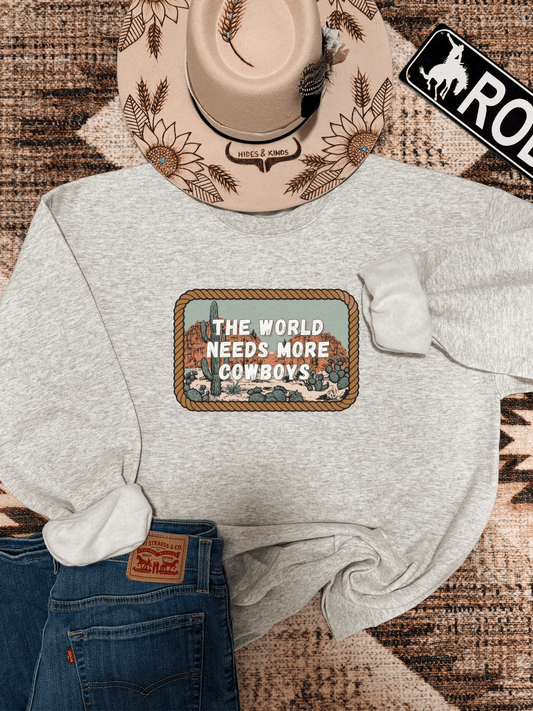 Heather grey crewneck laid flat with "The World Needs More Cowboys" graphic and desert cactus scene