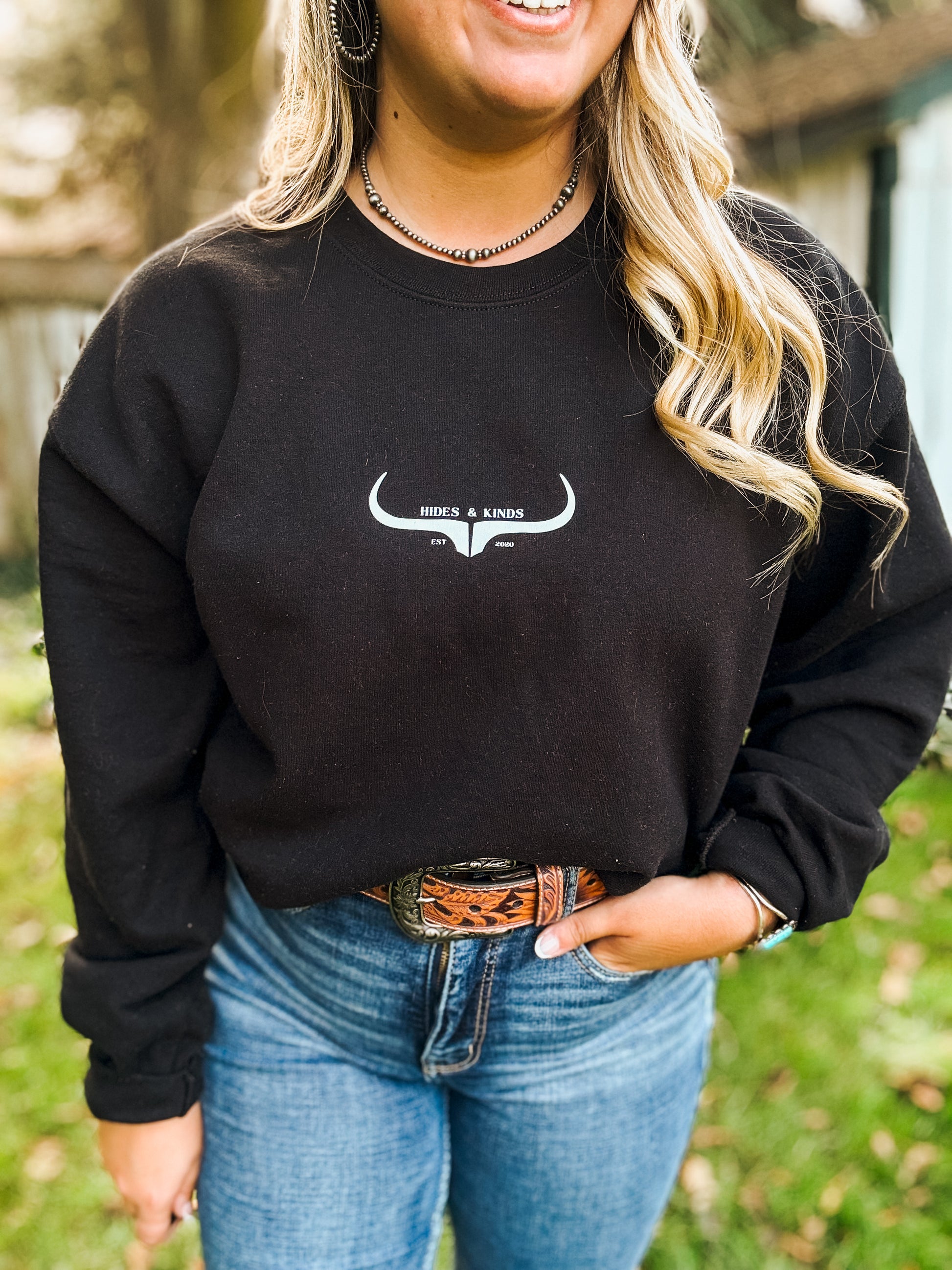 Woman wearing black Hides & Kinds crewneck with white longhorn logo, paired with a western tooled leather belt and jeans , casual outdoor lifestyle look.