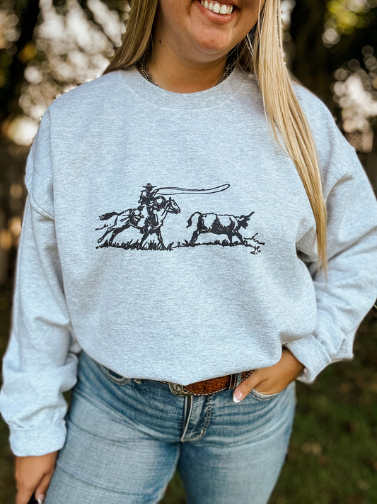 Heather grey western crewneck featuring an illustrated black roping scene of a cowboy on horseback chasing a steer. Worn tucked into denim with a tooled leather belt.