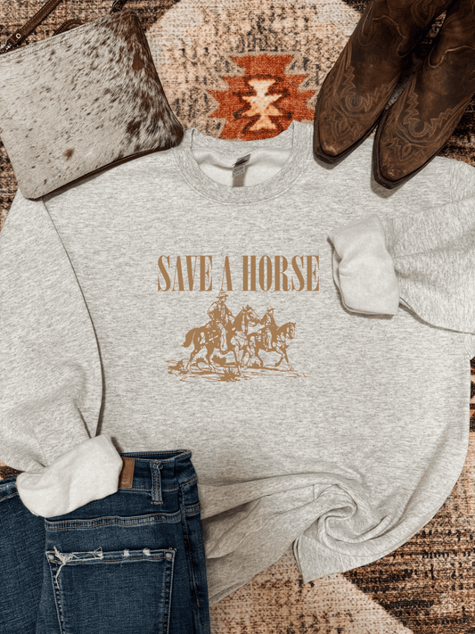 Save A Horse Crewneck in ash grey featuring cowboy riders in tan print, styled with denim, cowhide bag, and boots. Western graphic sweatshirt by Hides & Kinds.