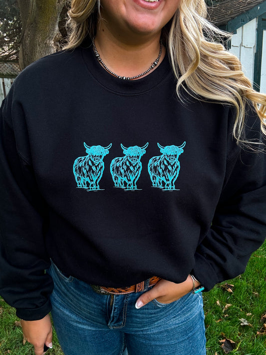 Close up view of a woman wearing the Hides & Kinds black crewneck with three turquoise highland cow graphics, styled with denim jeans and Western accessories, standing outdoors.