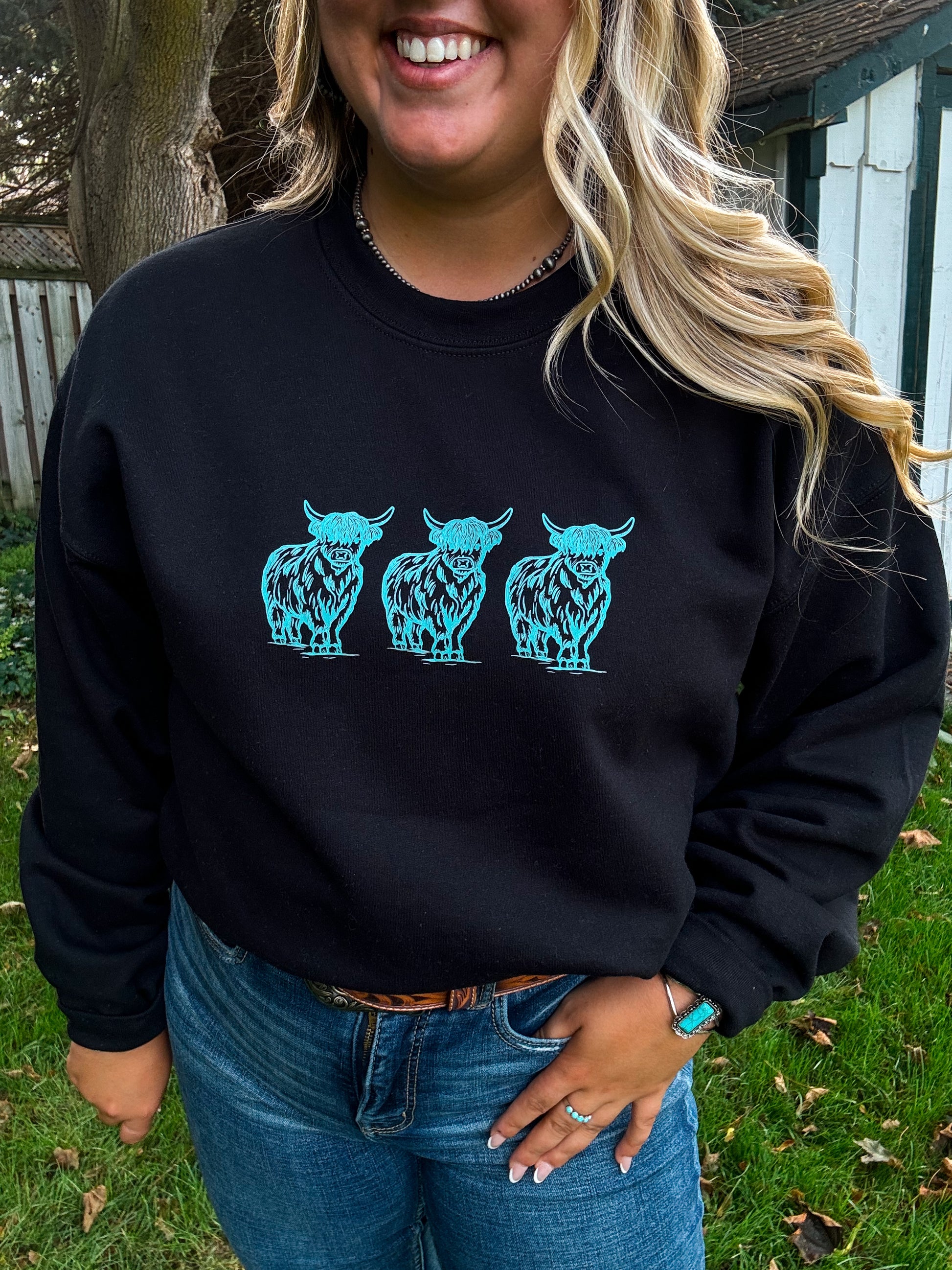 Smiling female model in the Hides & Kinds Turquoise Highland Crewneck, featuring bold turquoise cow designs, paired with western belt, turquoise jewelry, and a rustic background.