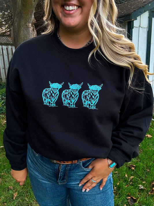 Smiling female model in the Hides & Kinds Turquoise Highland Crewneck, featuring bold turquoise cow designs, paired with western belt, turquoise jewelry, and a rustic background.