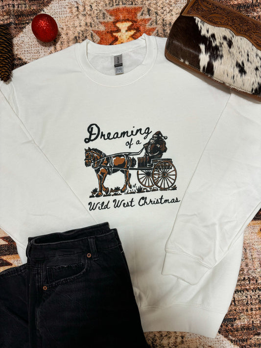 Western Christmas crewneck laid flat with festive pine cones, red ornaments, cowhide purse, and black denim jeans, cowboy Santa graphic with “Dreaming of a Wild West Christmas” text on a white sweatshirt