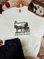 Western Christmas crewneck laid flat with festive pine cones, red ornaments, cowhide purse, and black denim jeans, cowboy Santa graphic with “Dreaming of a Wild West Christmas” text on a white sweatshirt