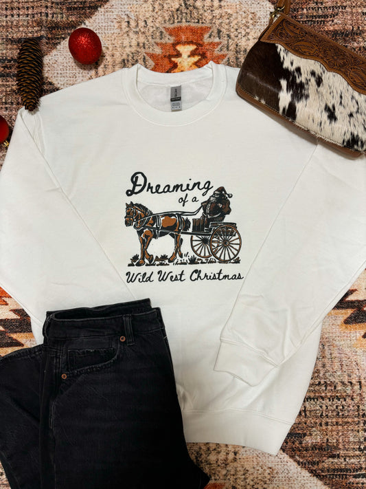 White crewneck featuring a vintage style cowboy Santa riding a horse-drawn wagon with “Dreaming of a Wild West Christmas” text, styled flat lay with black denim and rustic western holiday accents