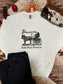 White crewneck featuring a vintage style cowboy Santa riding a horse-drawn wagon with “Dreaming of a Wild West Christmas” text, styled flat lay with black denim and rustic western holiday accents