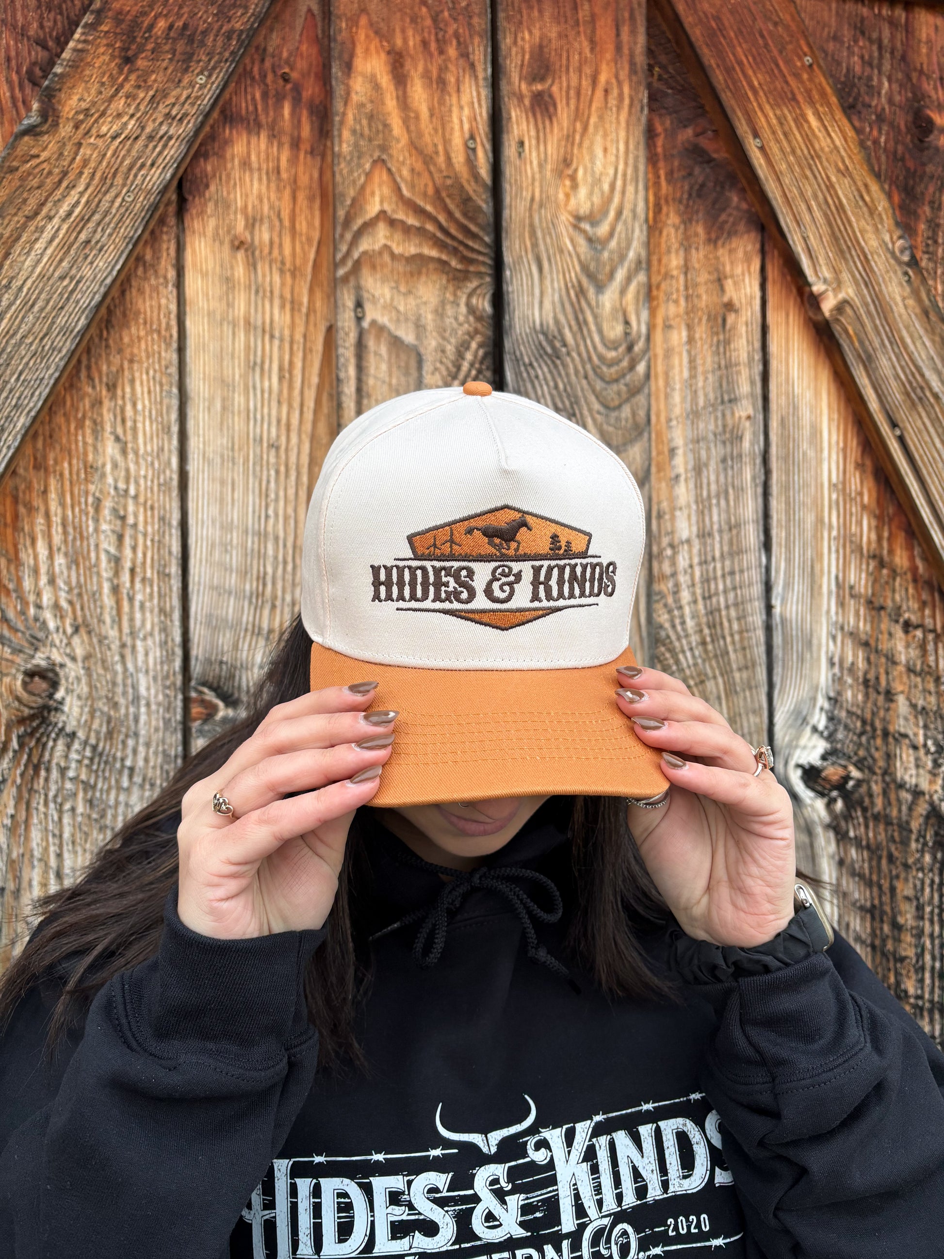 Front view of tan and cream Hides & Kinds Houston Hat styled on female model in front of rustic barn wall.