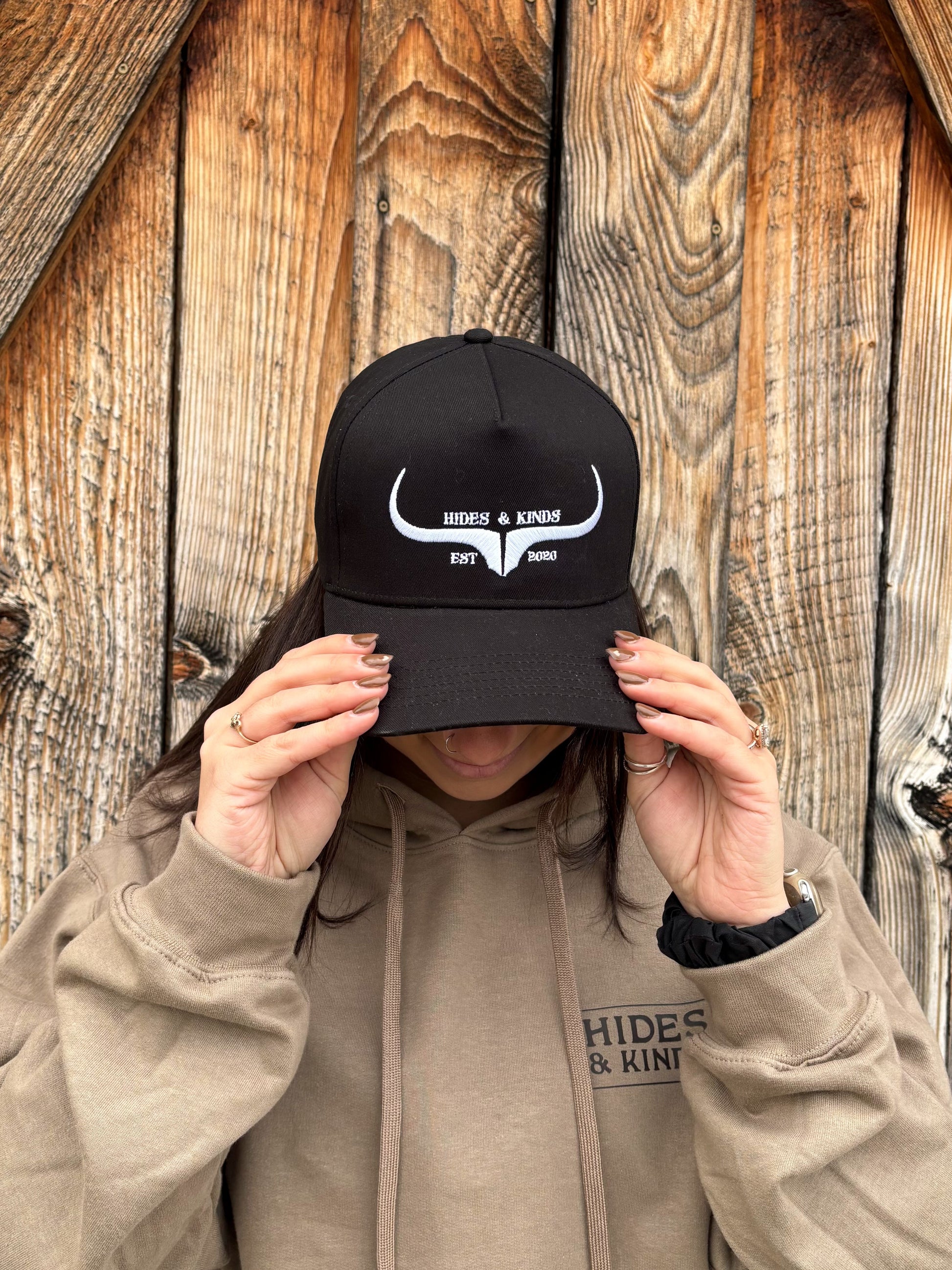 Front view of black and white Hides & Kinds Original Logo Hat styled on female model in front of rustic barn wall.