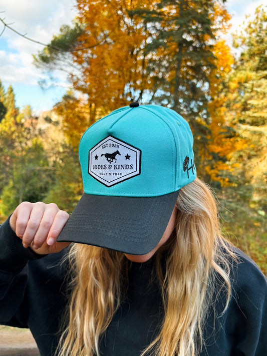 Side angle of teal and black Hides & Kinds Wild N Free Hat showing front logo patch.
