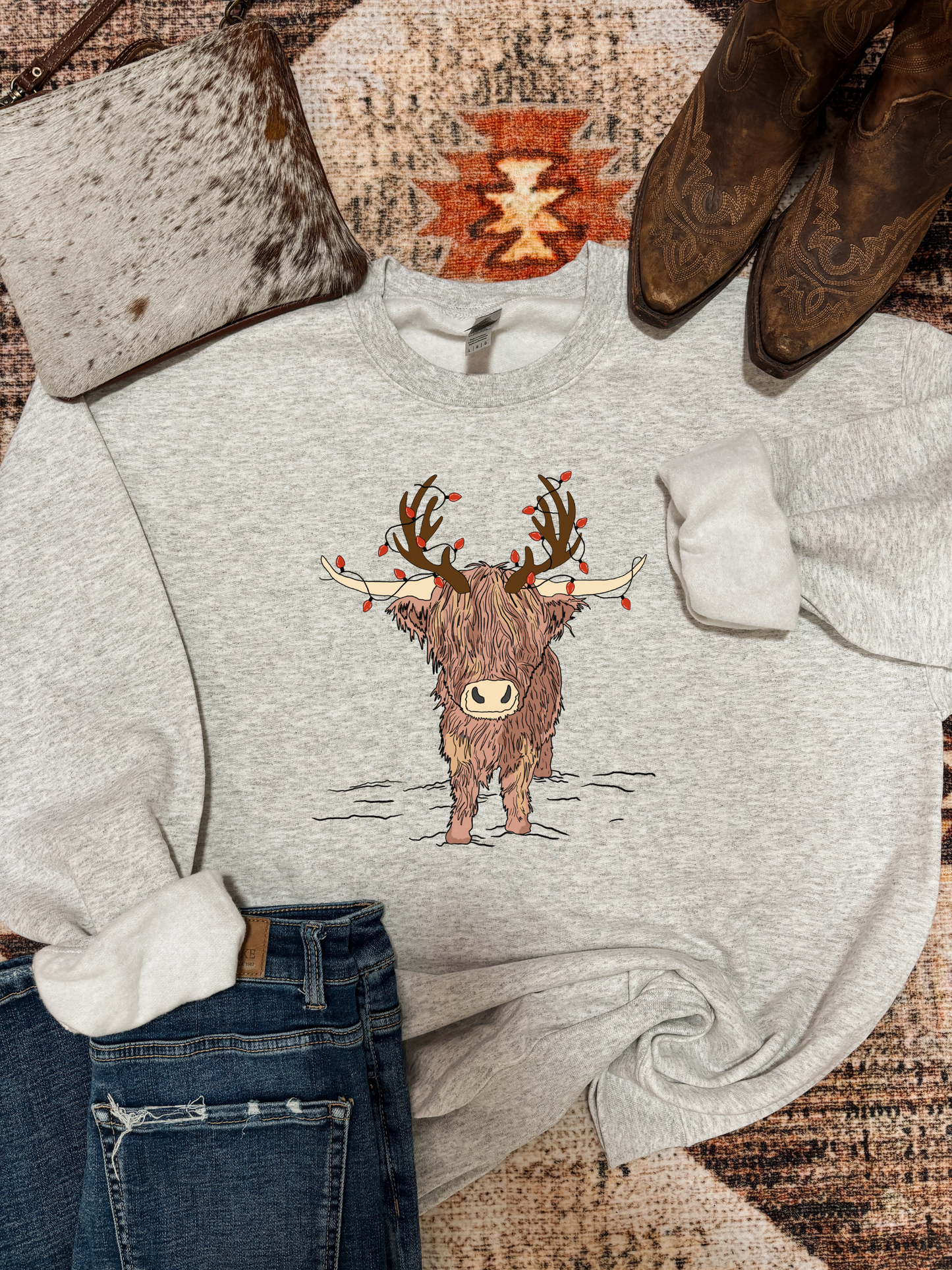 Ash grey Highland Christmas crewneck with festive highland cow design, styled with ripped jeans, brown cowboy boots, and a speckled cowhide clutch on a rustic western rug