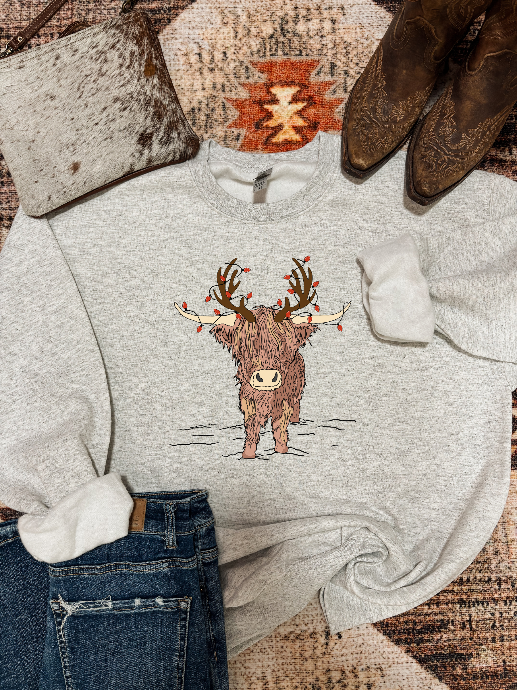 Ash grey Highland Christmas crewneck with festive highland cow design, styled with ripped jeans, brown cowboy boots, and a speckled cowhide clutch on a rustic western rug