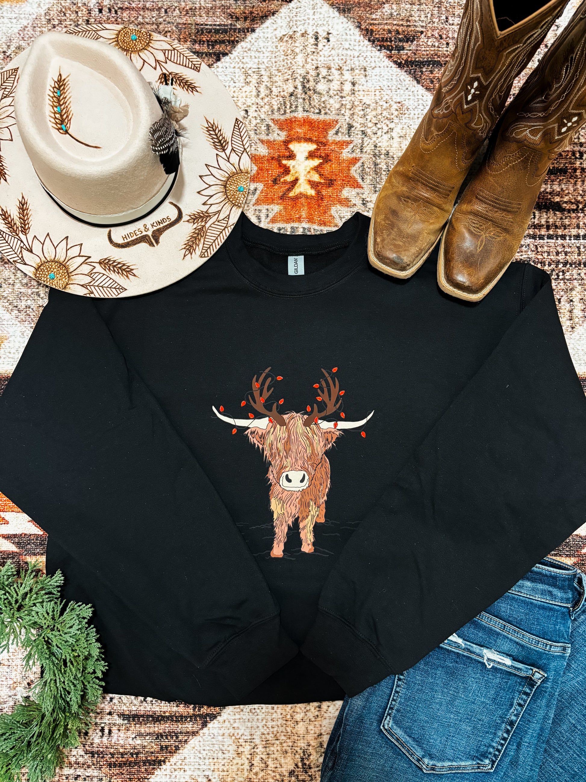Festive holiday crewneck with highland cow design laid out with jeans, cowgirl boots, and a hat with sunflower embroidery