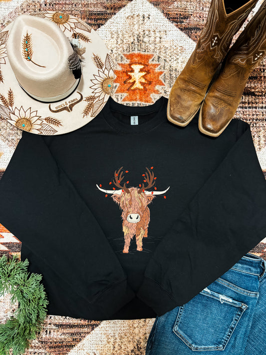 Festive holiday crewneck with highland cow design laid out with jeans, cowgirl boots, and a hat with sunflower embroidery