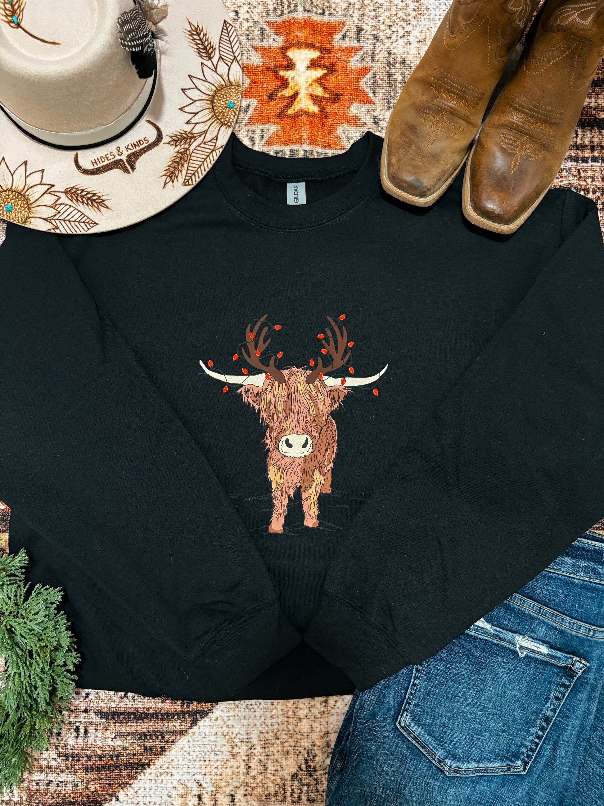 Black crewneck sweatshirt featuring a highland cow with antlers and red berries, styled with denim, boots, and a western hat