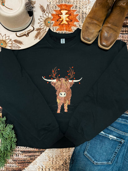 Black crewneck sweatshirt featuring a highland cow with antlers and red berries, styled with denim, boots, and a western hat