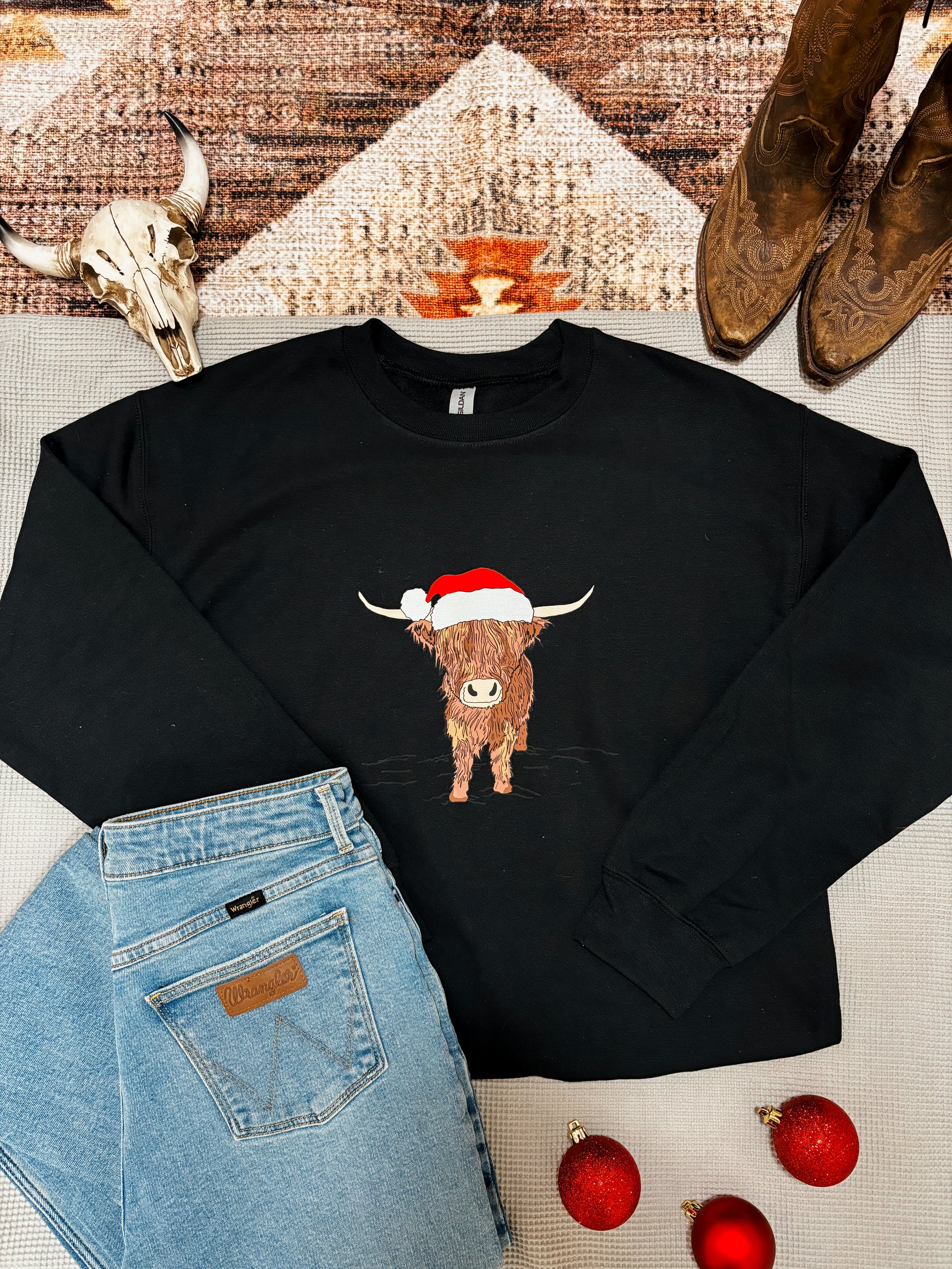 Western holiday sweatshirt with Highland cow design wearing Santa hat, styled with red ornaments and denim.