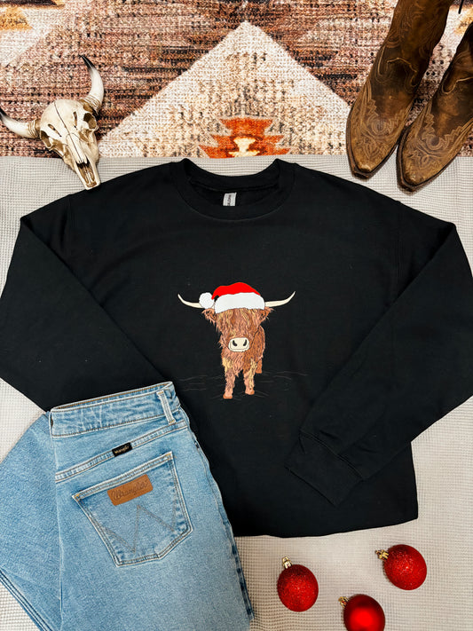 Western holiday sweatshirt with Highland cow design wearing Santa hat, styled with red ornaments and denim.
