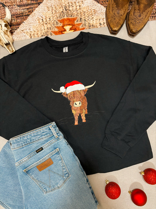 Flat lay of black Highland Santa crewneck with festive Highland cow in Santa hat, styled with jeans and boots.