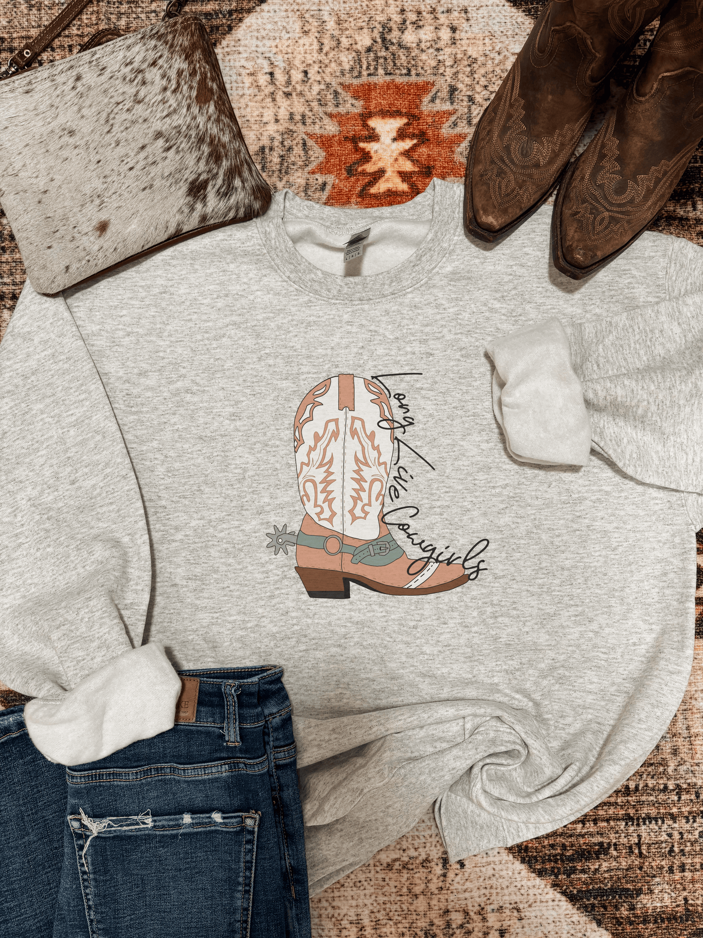 Ash grey “Long Live Cowgirls” graphic crewneck with western boot design, styled flat lay with jeans, cowhide clutch, and brown cowboy boots on a rustic southwestern rug