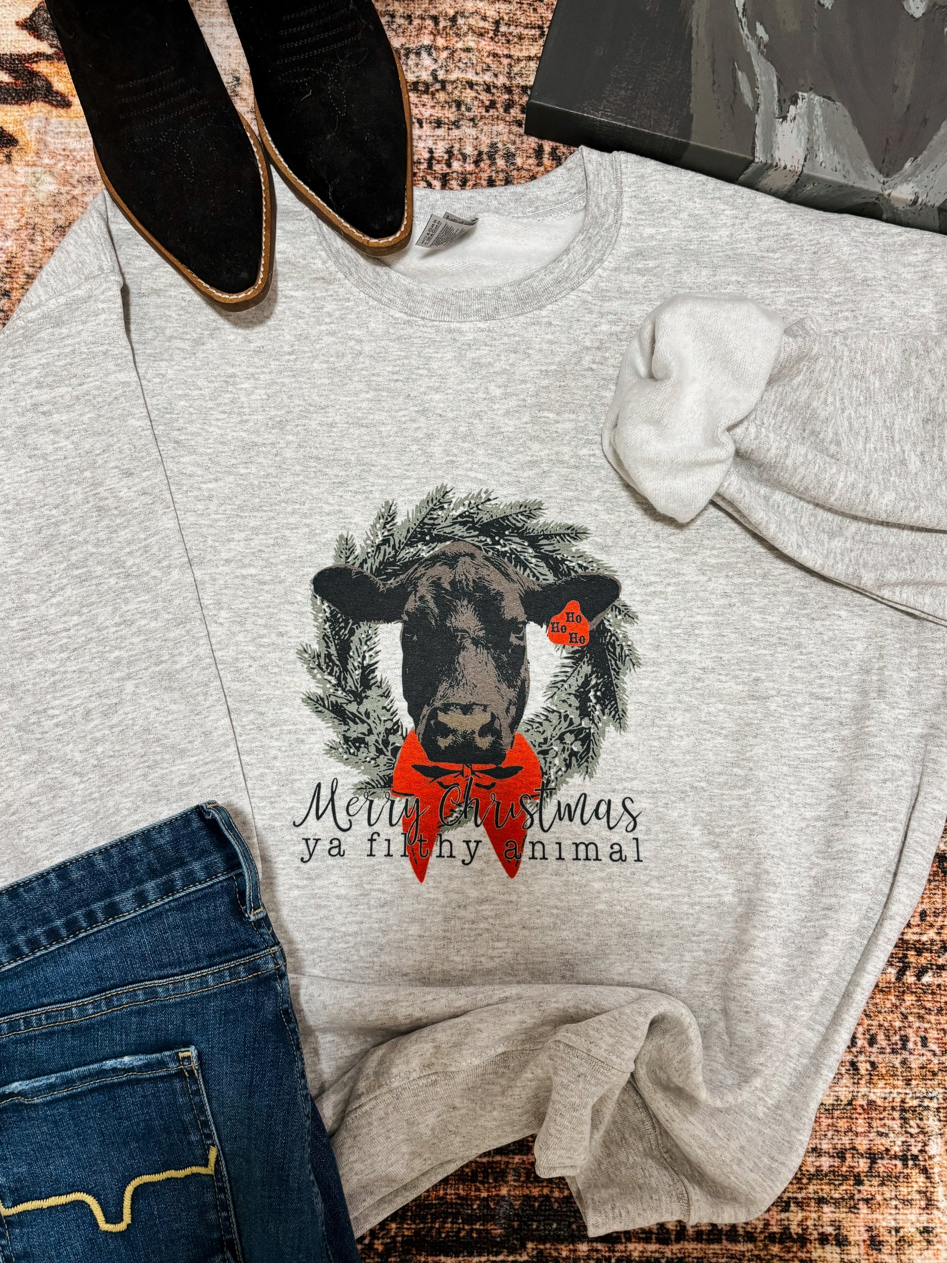 Flat lay of the Merry Christmas Ya Filthy Animal Crewneck with black suede boots and denim jeans, showcasing a festive cow graphic with a red bow and wreath