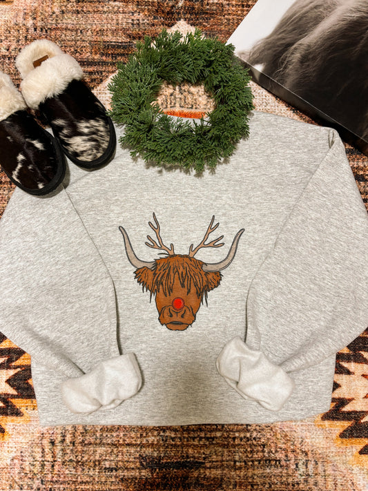 Flat lay of the "Moodolph Highland Crew" Christmas sweatshirt featuring a Highland cow with antlers and a red nose, laid on a western rug with cow print slippers, a mini pine wreath, and neutral cozy styling.