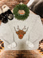 Flat lay of the "Moodolph Highland Crew" Christmas sweatshirt featuring a Highland cow with antlers and a red nose, laid on a western rug with cow print slippers, a mini pine wreath, and neutral cozy styling.