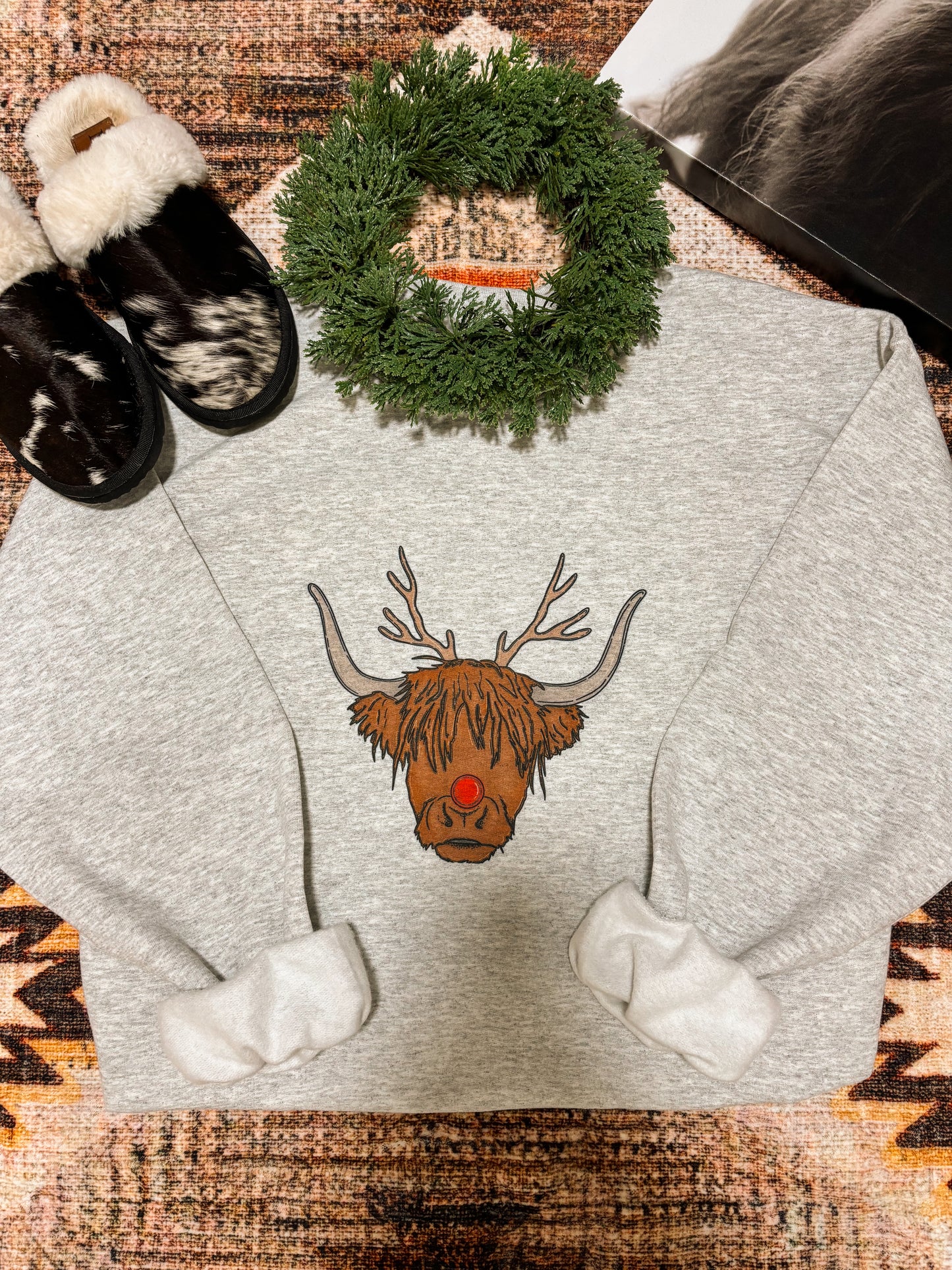 Detail shot of the Moodolph Highland Crewneck sweater showing the full front graphic, Highland cow as Rudolph, styled on a warm toned western rug with holiday props.