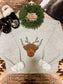 Detail shot of the Moodolph Highland Crewneck sweater showing the full front graphic, Highland cow as Rudolph, styled on a warm toned western rug with holiday props.