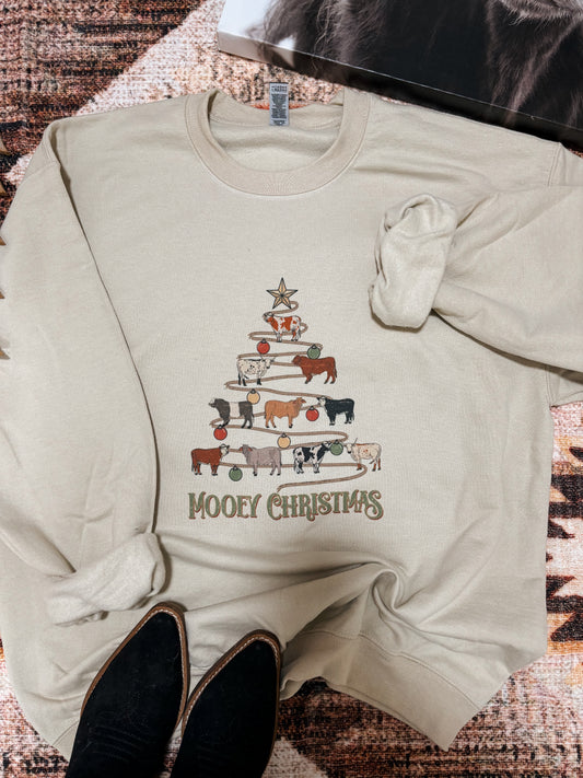 Sand coloured crewneck featuring a stacked cow graphic in the shape of a Christmas tree with festive ornaments and "Mooey Christmas" text, styled with black cowgirl boots on a rustic rug.