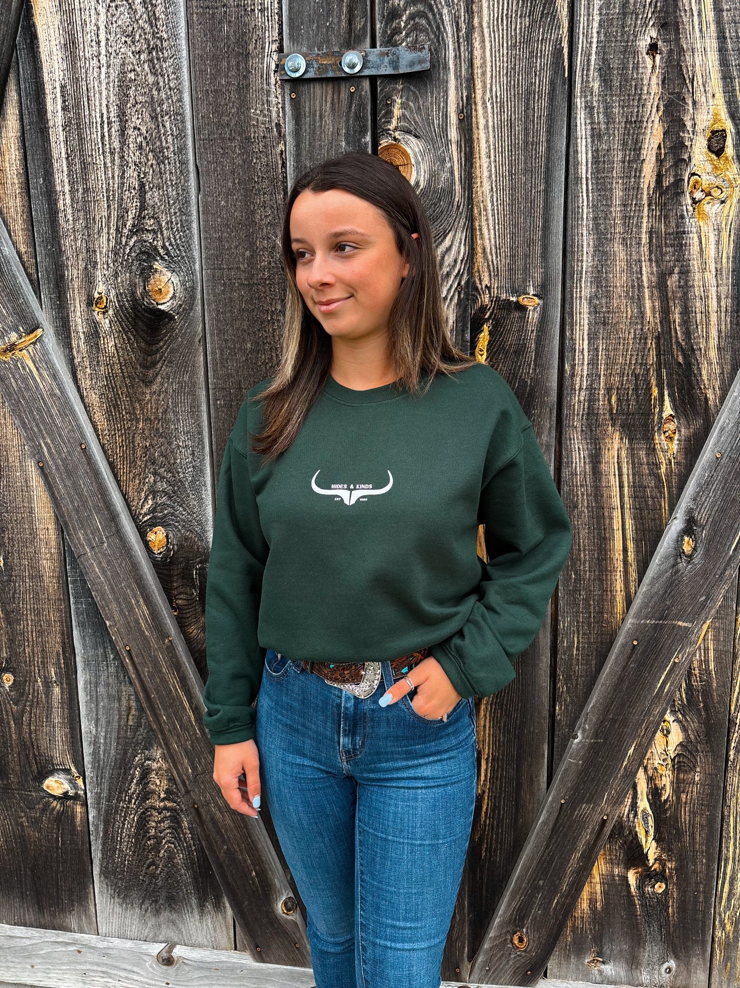 Woman wearing Hides & Kinds forest green crewneck with longhorn logo in front of wooden barn wall
