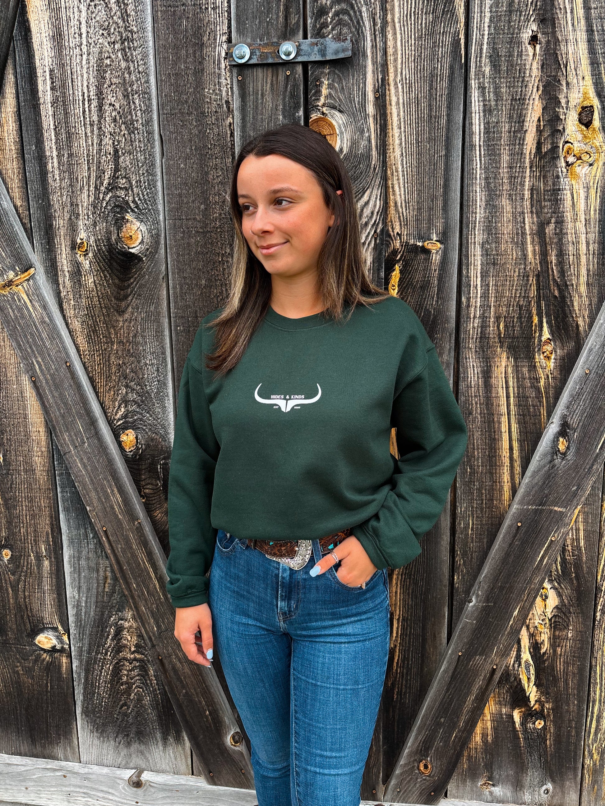 Woman wearing Hides & Kinds forest green crewneck with longhorn logo in front of wooden barn wall