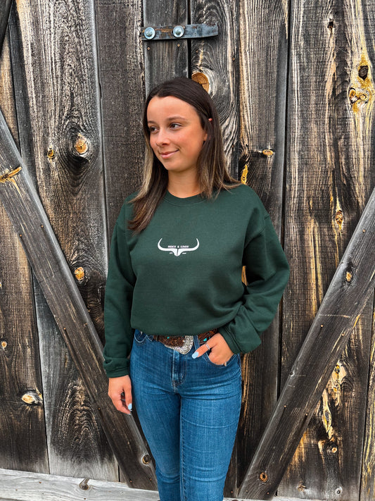 Woman wearing Hides & Kinds forest green crewneck with longhorn logo in front of wooden barn wall
